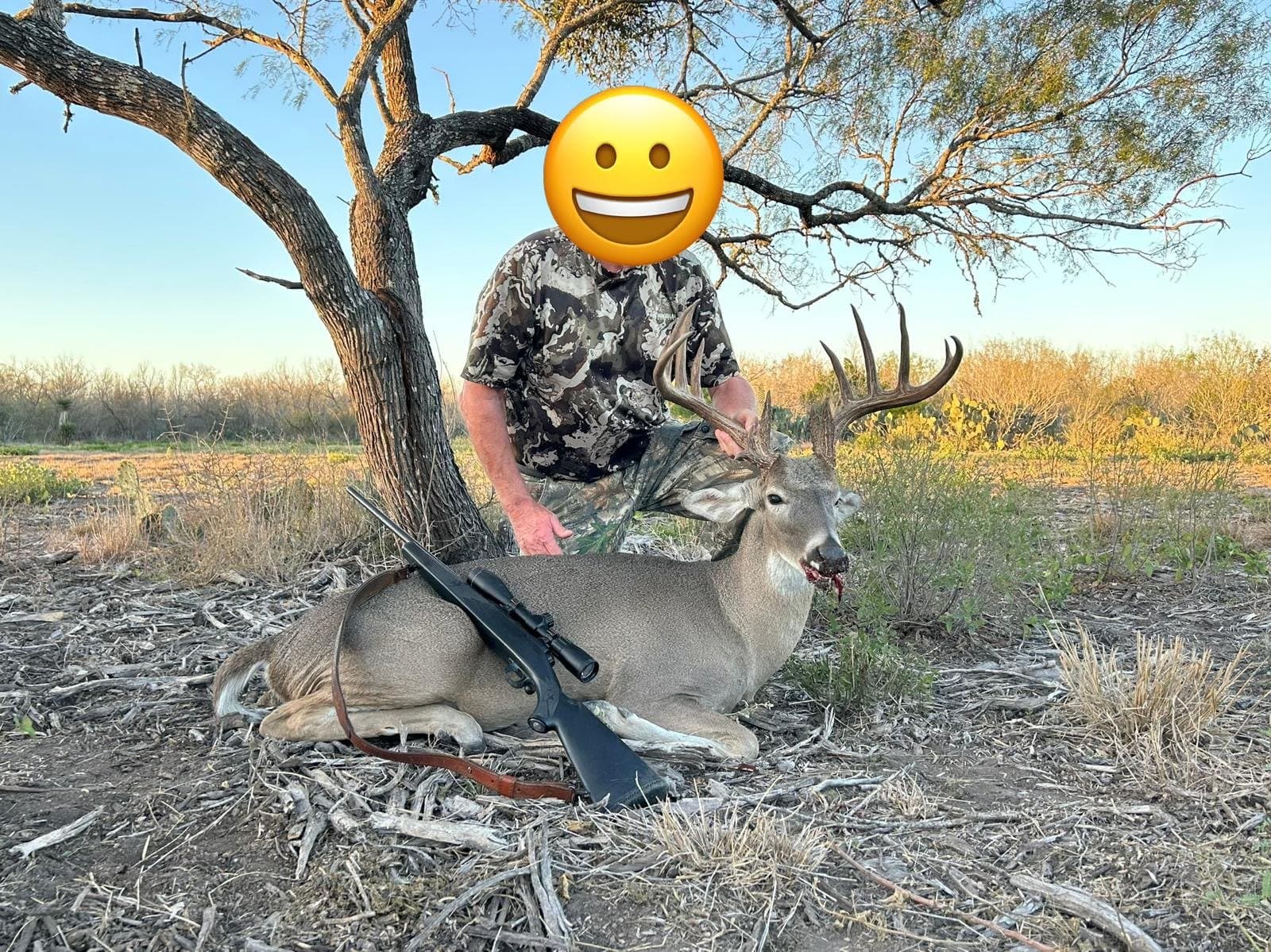 Hunter kneeling behind a harvested whitetail buck beneath a mesquite tree